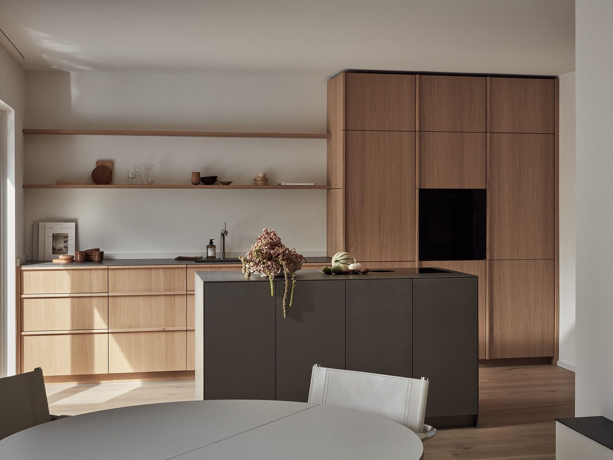 Eine moderne Küche mit hellen Holzschränken, einer dunkelgrauen Kücheninsel und minimalistischer Einrichtung. Das natürliche Licht unterstreicht die klaren Linien und schafft eine ruhige Atmosphäre