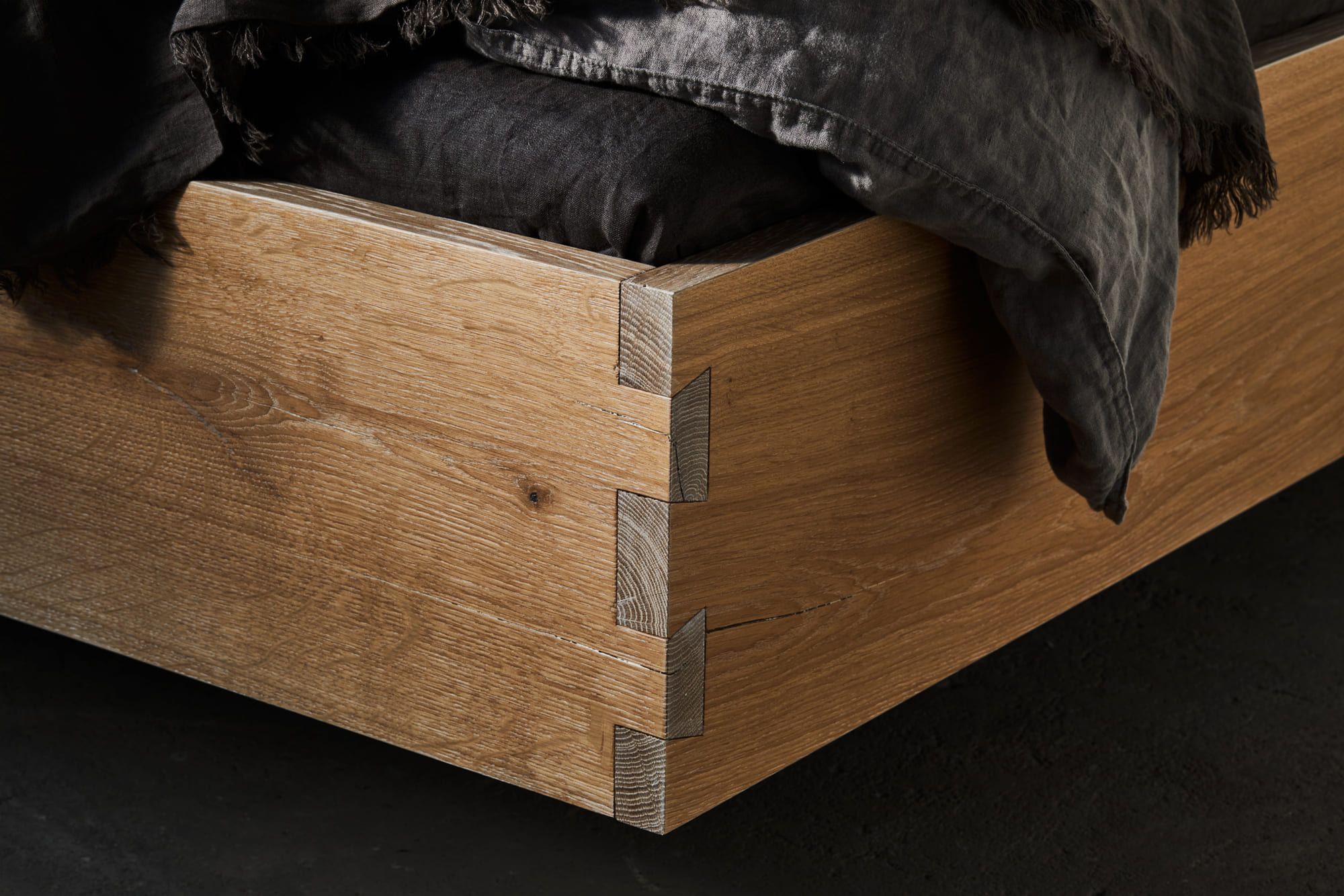 Nahaufnahme einer Ecke eines Holzbettes mit Schwalbenschwanzverbindungen, die die handwerkliche Kunstfertigkeit unterstreicht. Dunkle Bettwäsche fällt über die Bettkante