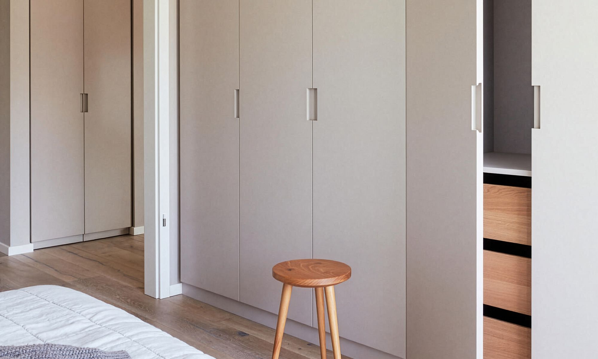 Ein minimalistisch eingerichtetes Schlafzimmer besticht durch beigefarbene Kleiderschränke mit schlichten Griffen, einen kleinen Holzhocker und einen offenen Schrank mit Holzschubladen