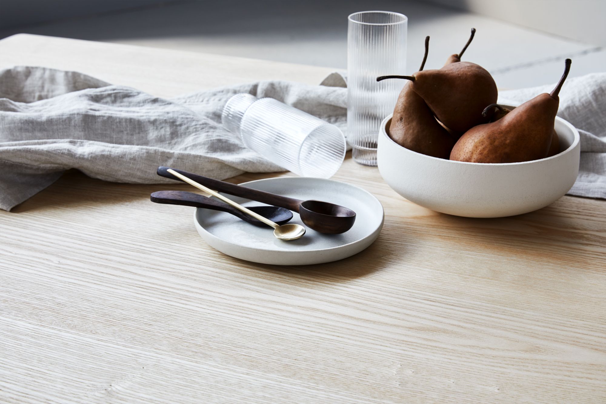 Eine minimalistische Tischdekoration mit einer weißen Schale voller Birnen, strukturierten Gläsern und einem Teller mit eleganten Löffeln
